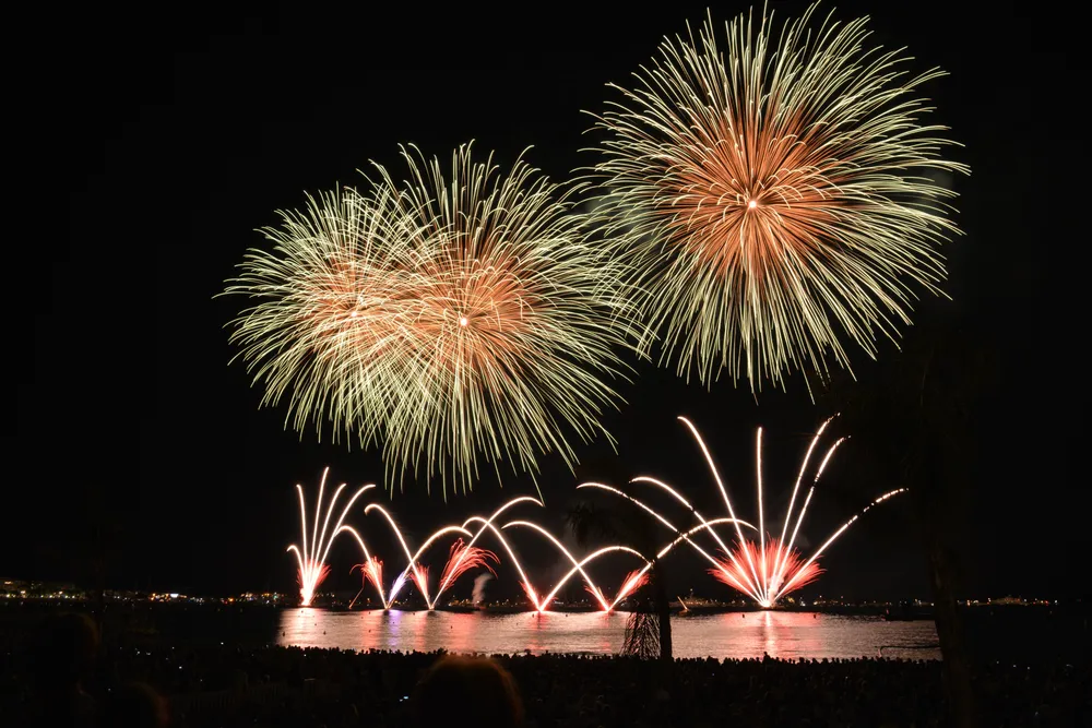 Spectaculaire feu d'artifice multicolore tiré au-dessus de la mer devant une plage bondée de monde la nuit
