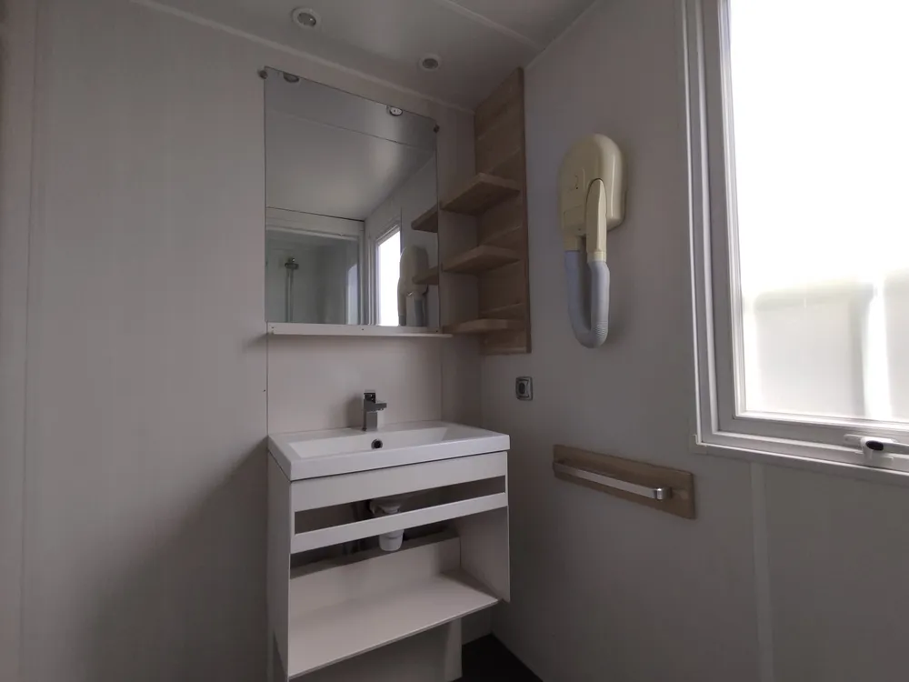 Salle d'eau d'un mobil-home avec lavabo, meuble de rangement, miroir et sèche-cheveux mural.