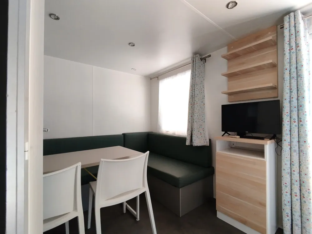 Coin salon intérieur d'un mobil-home avec banquette, table, chaises et télévision.