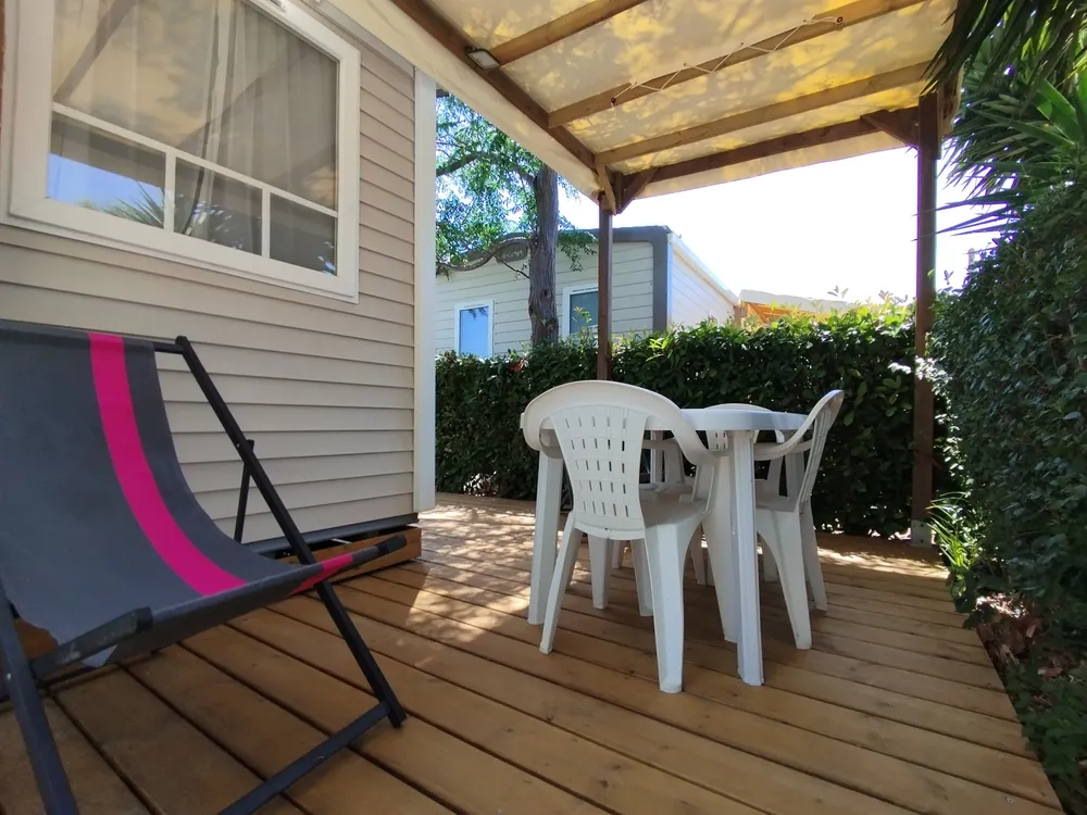 Terrasse en bois couverte d'un mobil-home avec table, chaises et chaise longue.