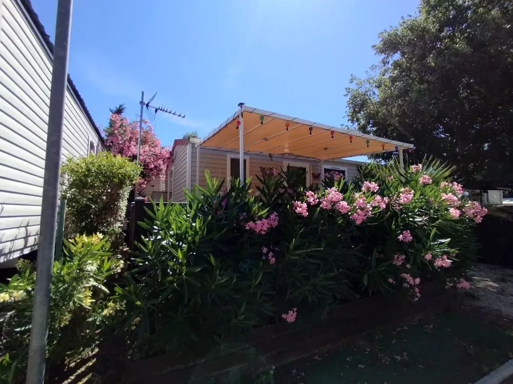 Mobil-home beige avec un grand auvent en toile et des lauriers-roses fleuris.