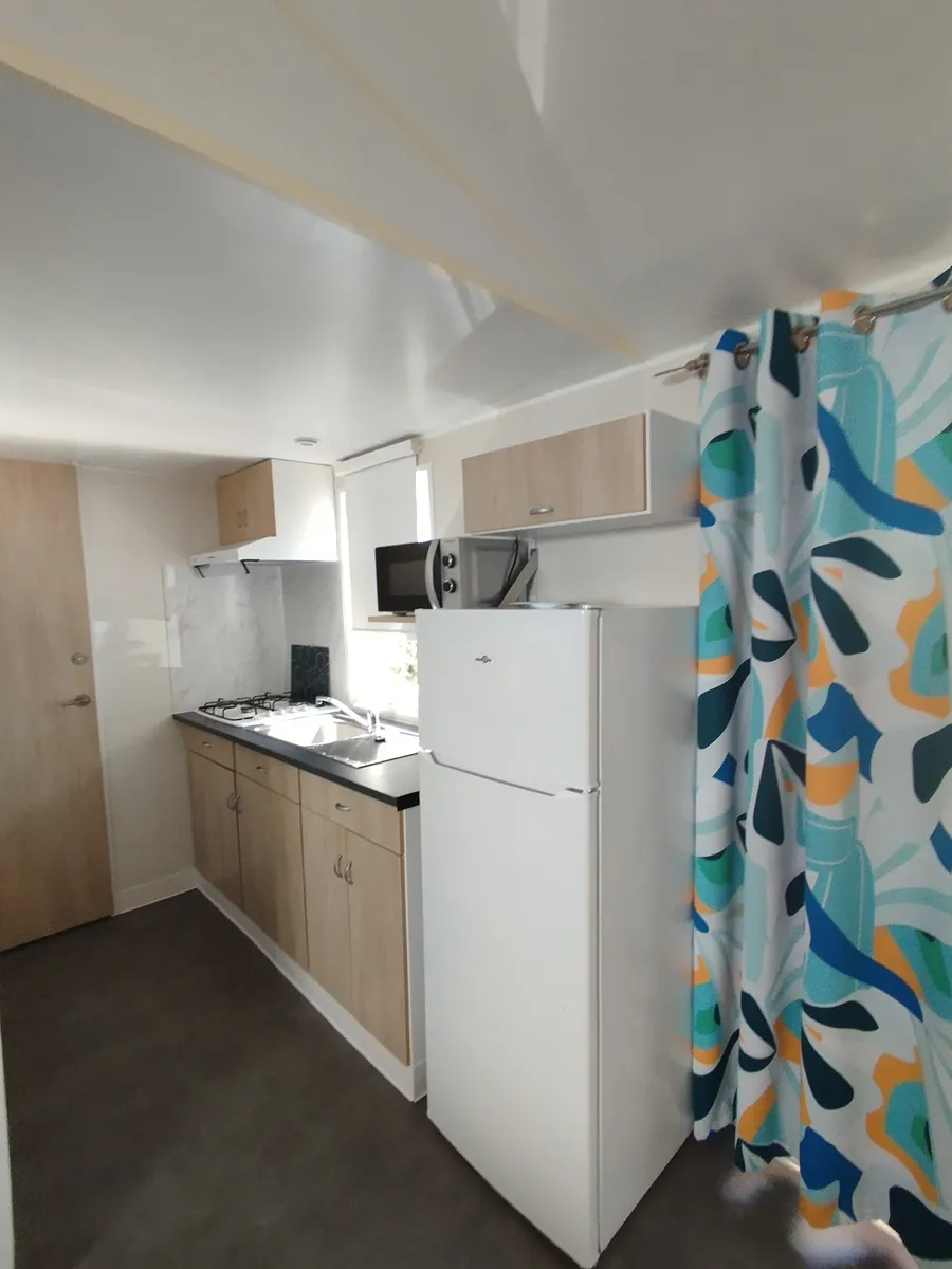 Vue en perspective de la cuisine linéaire d'un mobil-home avec un rideau décoratif coloré.