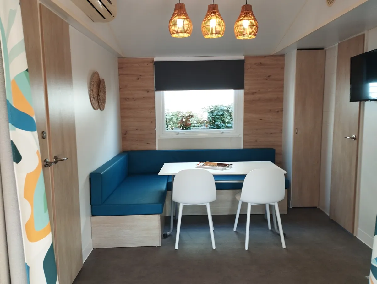 Espace de vie d'un mobil-home avec coin repas bleu, portes en bois clair et télévision fixée au mur