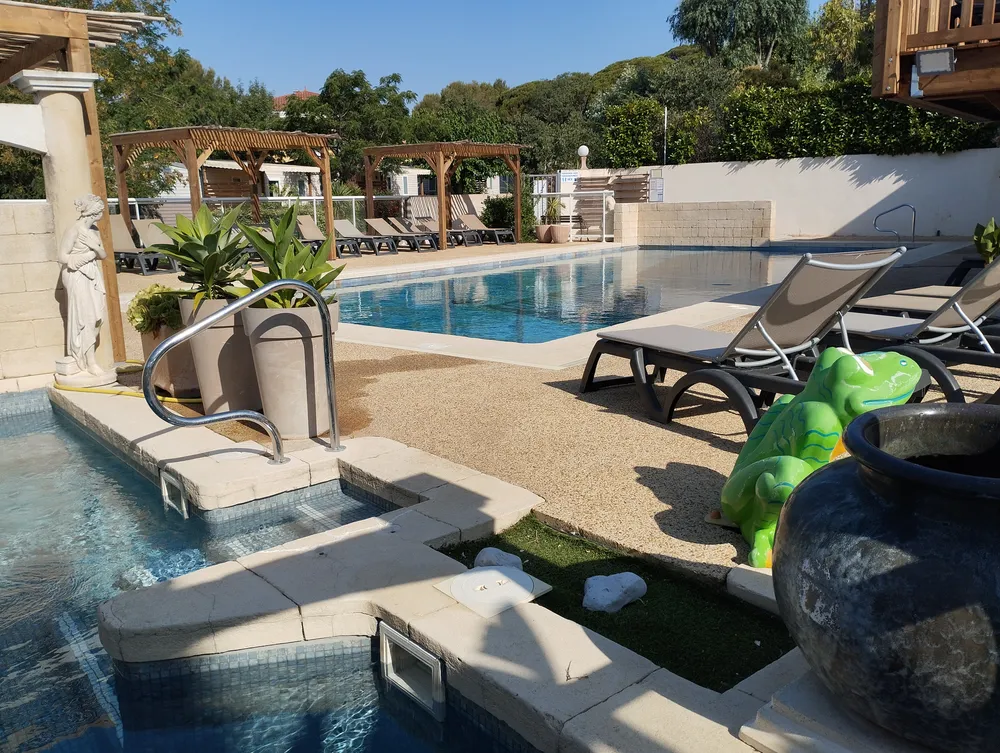 Espace aquatique de camping avec piscine, transats sous tonnelles en bois, plantes en pot et pataugeoire pour enfants en premier plan.