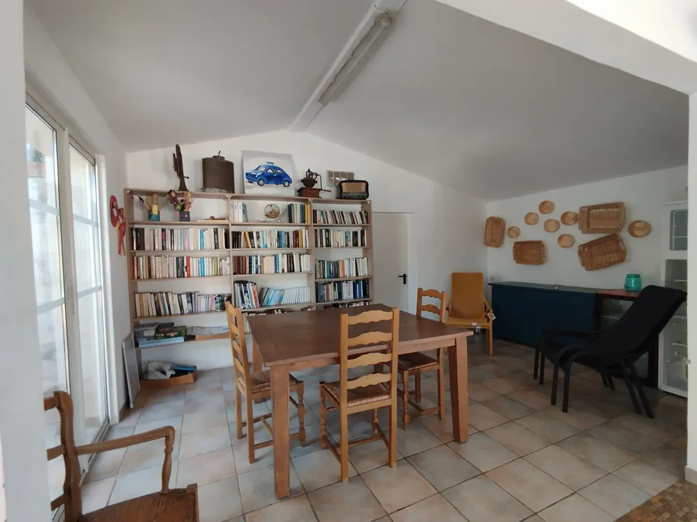Salle commune lumineuse avec une grande table en bois, des chaises en paille et une grande bibliothèque blanche remplie de livres.